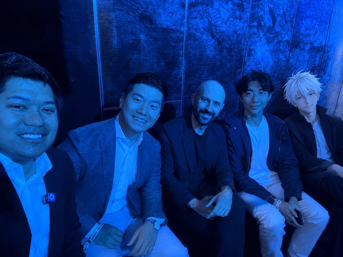 A group photo of five people sitting together in blue-lit surroundings, appearing to be at some kind of event or premiere, with a person in a white-haired anime-style costume on the far right.