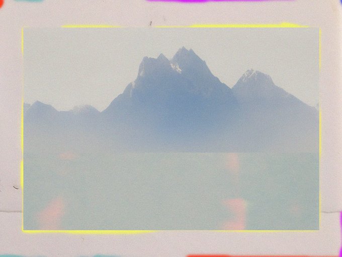 A stylized photo of jagged mountain peaks partially obscured by mist or low clouds, framed by a yellow border with a colorful, blurry film-leak effect.
