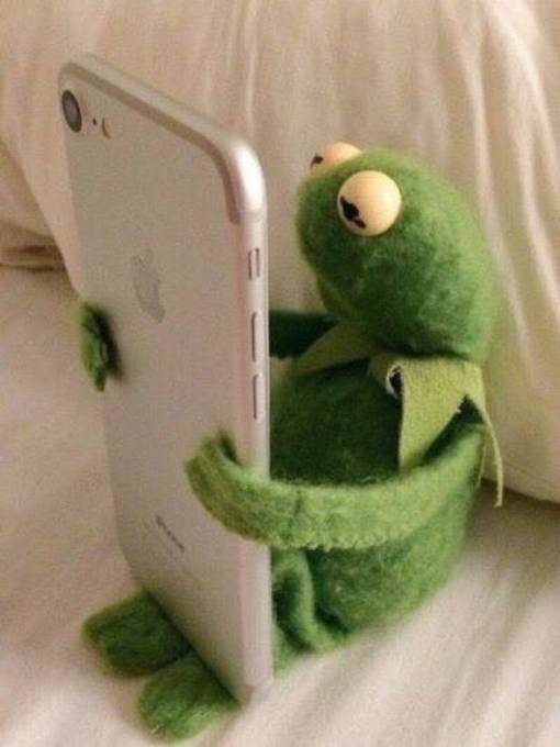 A green felt Kermit the Frog plush toy appears to be hugging or holding a silver iPhone from behind, sitting on a white fabric surface.