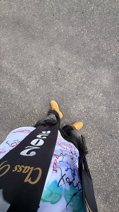 Graduating student stands on pavement wearing yellow boots, dressed in a white gown covered in colorful signatures, with a black "Class of 2026" sash reading "Class" visible — celebrating earning her BSC degree after a long, stressful journey.