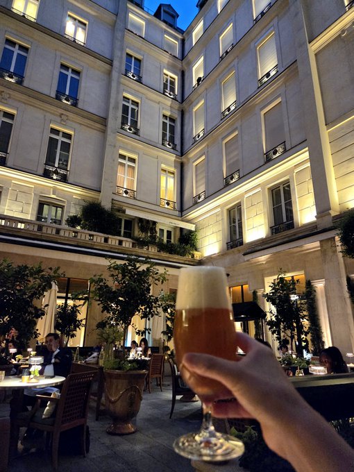 A hand raises a glass of beer in a toast at the elegant courtyard of the Park Hyatt Paris Vendôme at dusk, with the hotel's illuminated stone façade towering above. The image pairs with the caption's celebratory tone about ending a Europe stop and gearing up for the next leg of the trip.