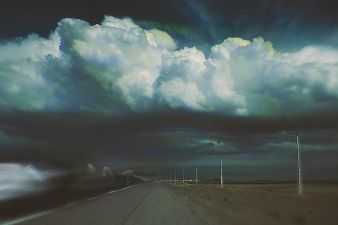 A dramatic landscape showing a dark road leading toward the horizon under massive, towering white and gray storm clouds, with telephone poles lining the right side of the road.