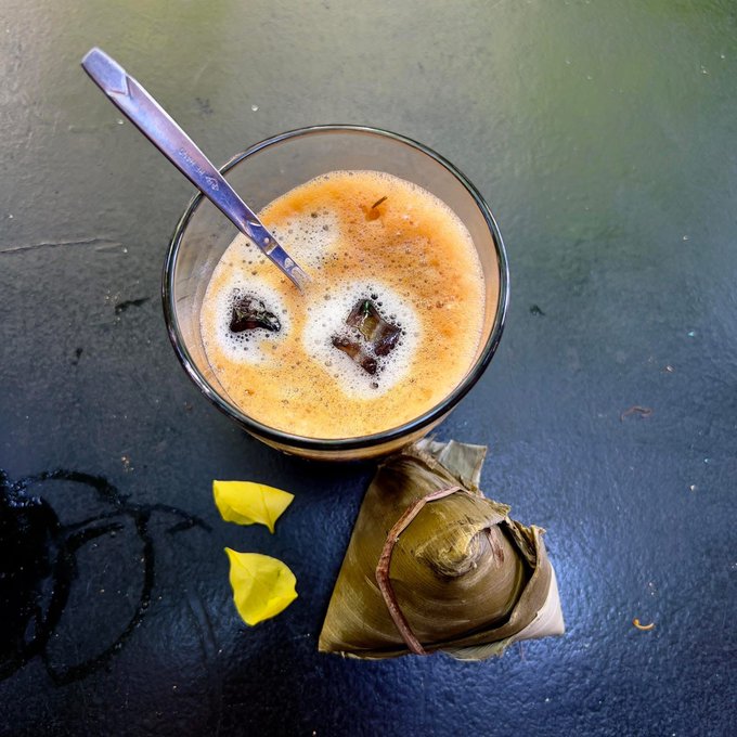 A top-down view of a glass of creamy, frothy beverage with ice cubes and a spoon, accompanied by a banana leaf-wrapped zongzi and yellow leaves on a dark surface.