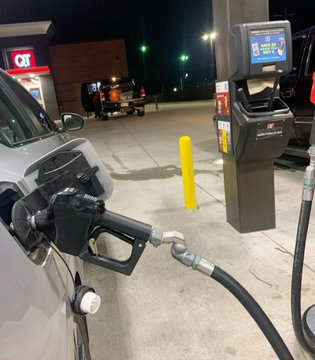 A nighttime photo at a QuikTrip (QT) gas station showing a fuel nozzle inserted into a vehicle's gas tank with the payment kiosk visible in the background.
