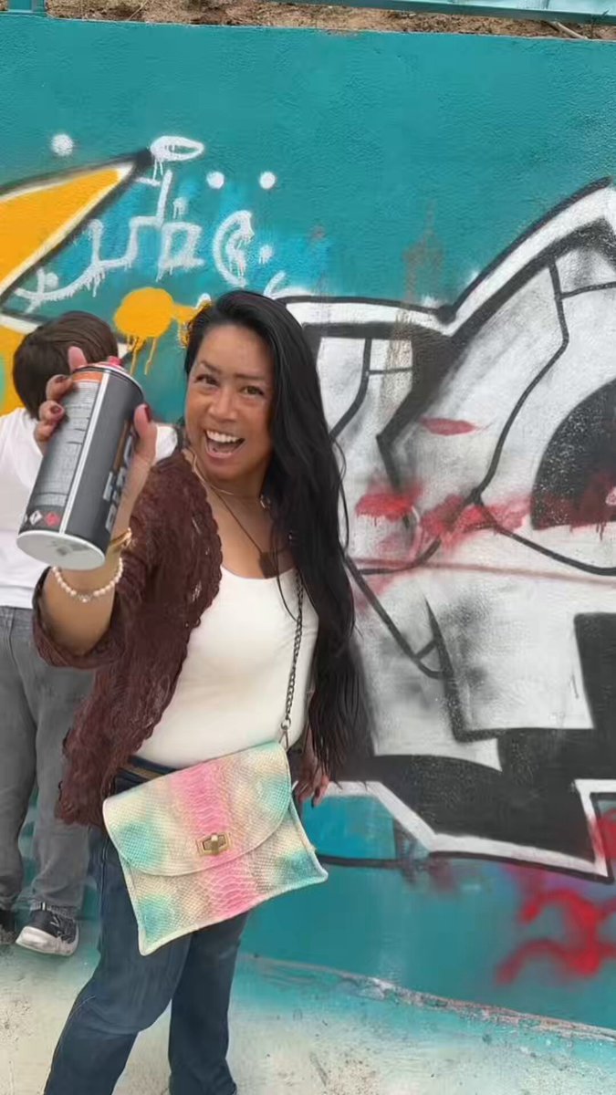 The image features a woman standing next to a colorful graffiti wall, holding up her hand with a spray paint can in front of her face. She appears to be posing for a photo or possibly creating art herself. The background includes a blue wall adorned with white graffiti and a yellow wall featuring a black and white design. There is also another person partially visible on the left side of the image, but they are not the main focus of the scene.