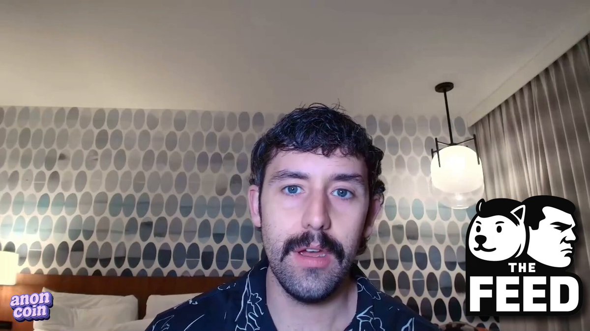 The image features a man with a beard wearing a black shirt, sitting at a desk or bed. He has his face turned towards the camera, giving an impression of being engaged in conversation or participating in a live stream. The background includes a wall adorned with blue and white circles, which could be interpreted as wallpaper or a pattern on the room's walls. The image also contains text that reads "THE FEED", possibly indicating the name of the post where the man is located or the topic being discussed.