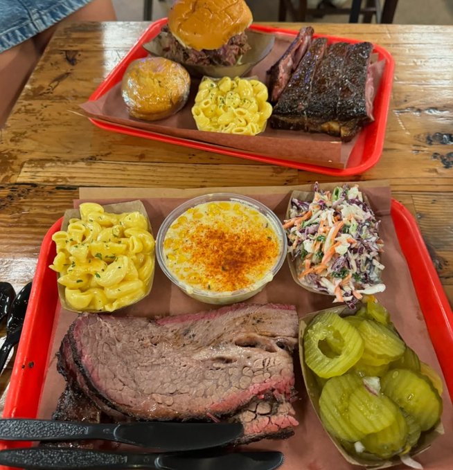 Two red trays loaded with Texas-style BBQ, including slices of smoked brisket and ribs, pulled pork sandwiches, mac and cheese, coleslaw, pickles, and a creamy seasoned side, served on butcher paper over a rustic wooden table. The hearty feast appears to be the group's way of celebrating after posting "gm" — fueling up on classic comfort food to match the bullish crypto energy.