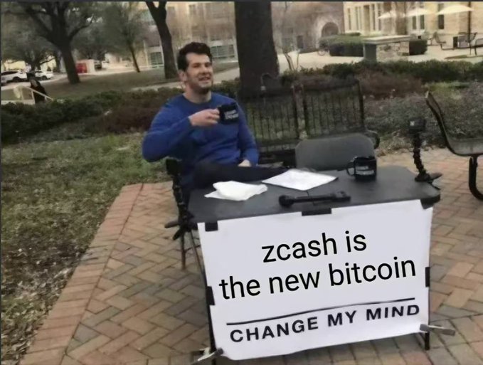 A man sitting outdoors at a table with a 'CHANGE MY MIND' sign that reads 'zcash is the new bitcoin'.