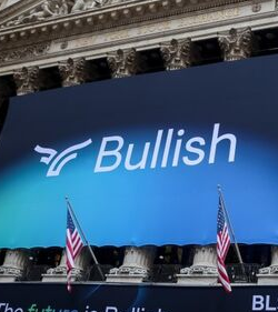 A banner reading "Bullish" with its logo is displayed in front of a classical building with columns, flanked by American flags, indicating a financial or corporate setting related to the cryptocurrency company Bullish. The image visually reinforces the post's news about Bullish's $1.28 billion Bitcoin holdings and its corporate identity, tied to its parent company Block.one and its ownership of CoinDesk.