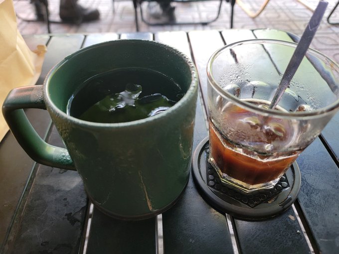 A green ceramic mug of dark liquid sits next to a glass of iced coffee on a black slatted table at an outdoor café.