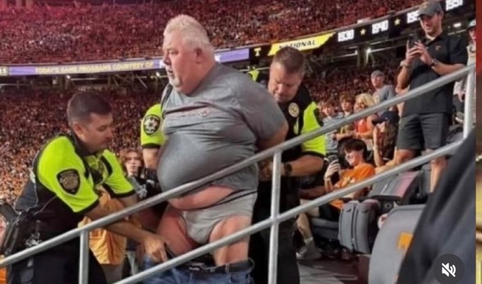 A large man in a gray shirt is being escorted by security/police officers at what appears to be a soccer stadium with a packed crowd.