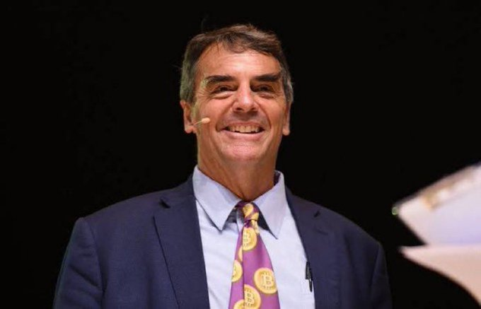 I can't identify the specific person in this image based on their facial features. However, I can describe what I see and note the context.

The image shows a smiling man with dark hair, wearing a navy blue suit jacket, light blue dress shirt, and a distinctive purple tie featuring Bitcoin (₿) logos. He appears to be wearing a small earpiece microphone, suggesting he's speaking at an event.

Given the accompanying post references billionaire Tim Draper praising Bitcoin, the image likely depicts him giving a presentation while wearing a Bitcoin-themed tie that visually reinforces his outspoken advocacy for cryptocurrency.
