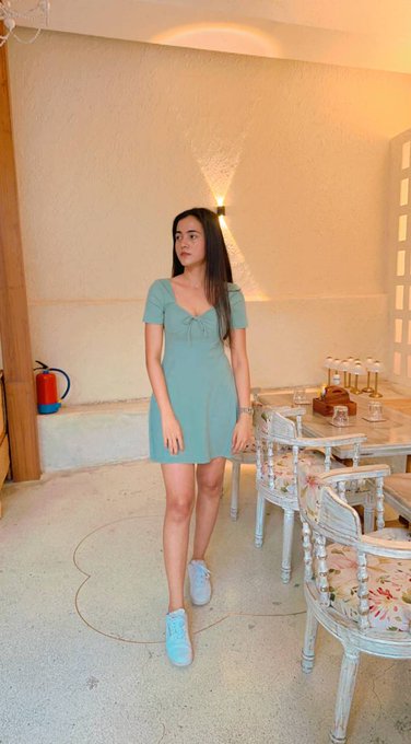 A young woman stands indoors wearing a light blue short-sleeved dress and white sneakers, posed next to a white ornate dining table and chairs.