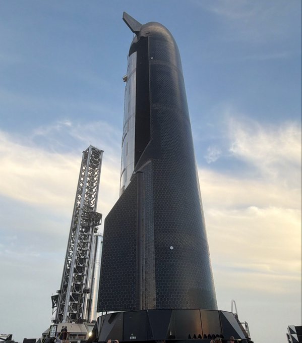 A tall black SpaceX Starship rocket stands vertically on a launch pad next to a metallic tower under a partly cloudy sky.