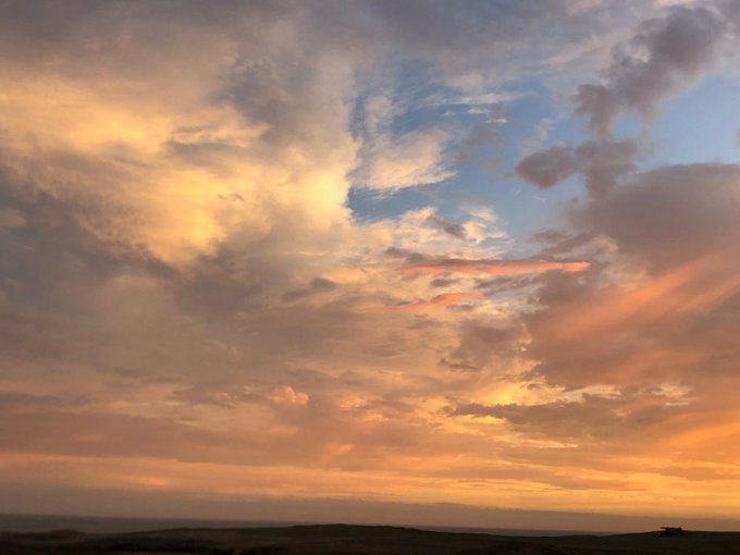 A scenic view of a dramatic sky at sunset or sunrise, featuring clouds illuminated in warm orange and golden hues against patches of blue, over a dark horizon.