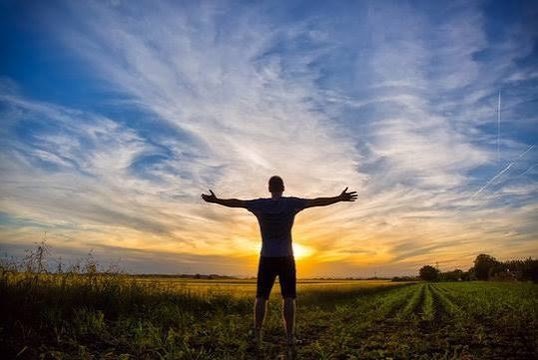 A silhouette of a person standing with arms outstretched in a field, facing a vibrant sunset under a cloudy sky.