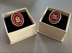 The image features two identical gold-colored pins, each adorned with a red circle design and the words "I'm a @MadLads x @Backpack believer" written on them. The pins are placed inside a white box or a small wooden case, which is positioned in front of the viewer. The background appears to be a gray surface, providing a neutral backdrop that allows the pins to stand out prominently.