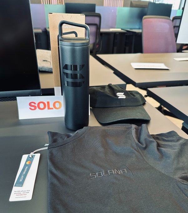 A photo of promotional merchandise on a conference table, including a black insulated tumbler with an Ethereum logo, a black Solana t-shirt with tag, a black cap, and signage reading "SOLO."