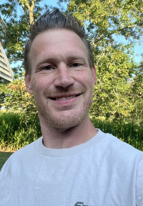 A selfie of a smiling man with short brown hair, wearing a white t-shirt, with trees and a house visible in the background.
