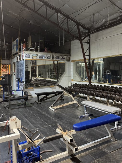 An interior view of a vintage gym featuring weight benches, dumbbell racks, weightlifting equipment, and mirrored walls under an industrial ceiling.