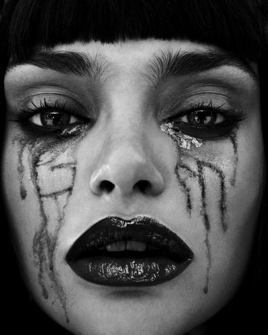 A black and white close-up portrait of a woman with dark bangs, dark lipstick, and dramatic makeup with dark streaks running down her cheeks from beneath her eyes.