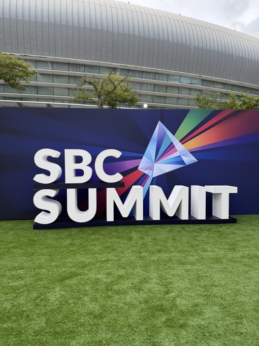 A large outdoor sign displaying "SBC SUMMIT" in bold white letters with a colorful star logo, set up on artificial turf in front of a modern convention center building.