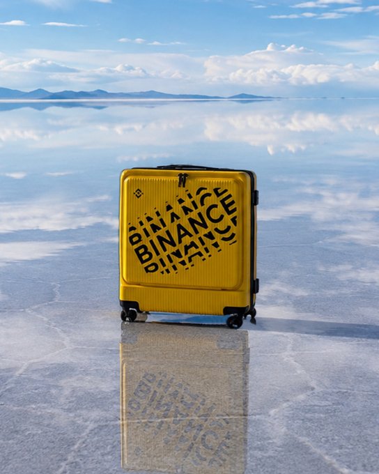 A yellow suitcase with "BINANCE" branding printed on it sits on the mirror-like surface of the Salar de Uyuni salt flat in Bolivia, reflecting the sky and clouds. The image ties into the post's caption about passing through Bolivia, juxtaposing a travel scene with the cryptocurrency exchange's branding in an unexpectedly scenic location.