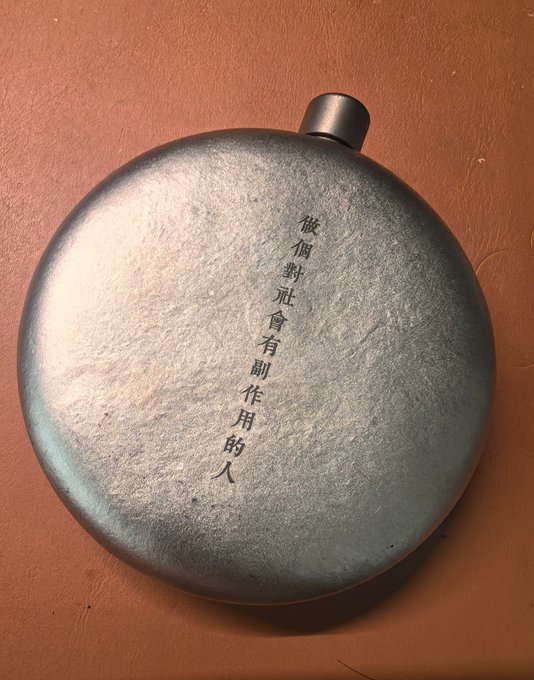 A metallic flask engraved with Chinese characters '被傾訴會有副作用的人' resting on a brown surface.