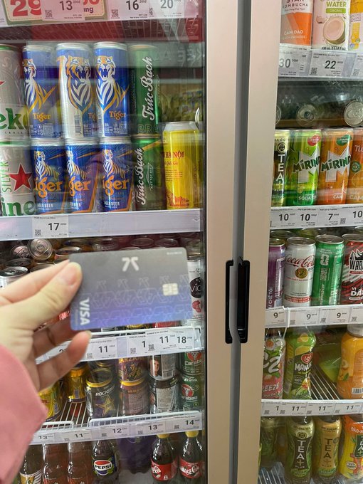 A hand holds a Visa credit card in front of a refrigerated display case stocked with Tiger, Mirinda, Coca-Cola, Sprite, and other assorted beverages.