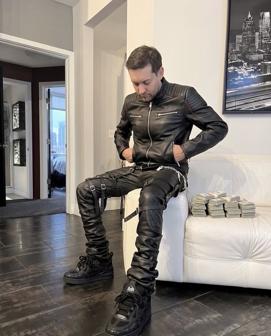 A man sits on a white leather couch wearing a full black leather outfit with harness straps and chains, next to stacks of cash, in a modern room setting.