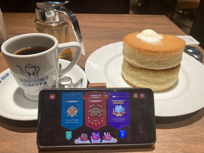 Hoshino Coffee table setting featuring a cup of coffee, a syrup dispenser, and a tall stack of fluffy pancakes with butter, with a phone displaying a mobile game in the foreground.