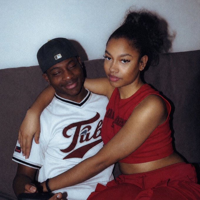 A photo of a young couple sitting together on a couch; the man wears a white Title jersey and baseball cap, while the woman wears a red crop top and pants.
