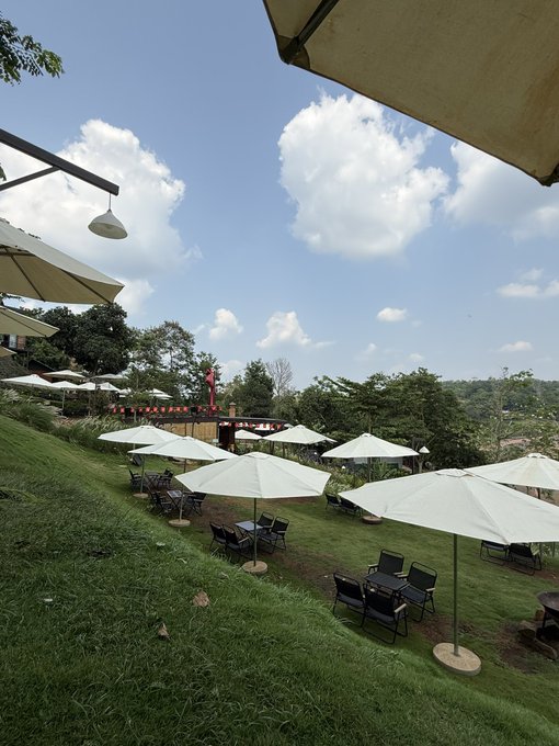 An outdoor hillside café/restaurant features rows of white umbrellas shading tables and chairs on a grassy lawn under a partly cloudy sky. The scenic setting complements the morning greeting post from @quipnetwork, suggesting a peaceful breakfast or brunch spot with a beautiful view.