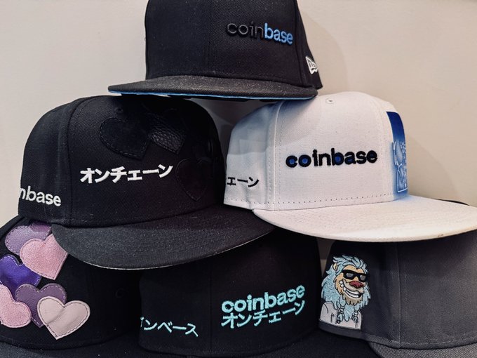 A display of stacked baseball caps featuring the Coinbase logo in English and Japanese (オンチェーン) text, with one cap showing a llama meme character.