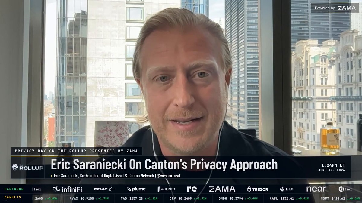 In the image, a man wearing a black shirt is sitting at his desk with a window behind him that offers an urban cityscape view. The man appears to be smiling for the camera while talking about privacy and governance participation in Canton. There are several other people visible in the background of the image, but they do not seem to be directly involved in the conversation.
