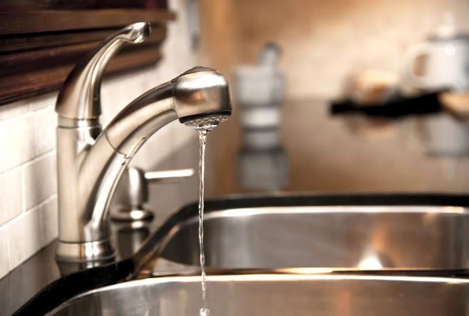 A sleek stainless steel kitchen faucet pours a steady stream of water into a polished sink, with a blurred countertop of containers in the background. The image ties to the post's lighthearted "the faucets in guys" caption by highlighting an everyday household fixture.