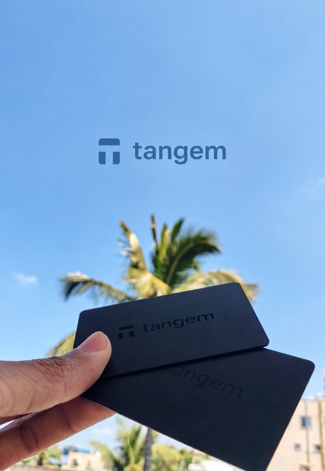 A promotional image showing a hand holding two black "tangem" crypto hardware wallet cards against a tropical beach background with palm trees and blue sky, with the tangem logo displayed in the upper portion.