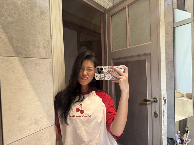 A woman takes a mirror selfie wearing a white and red 'Red Hot & Raw' cherry shirt, holding a phone with a floral case in a doorway.