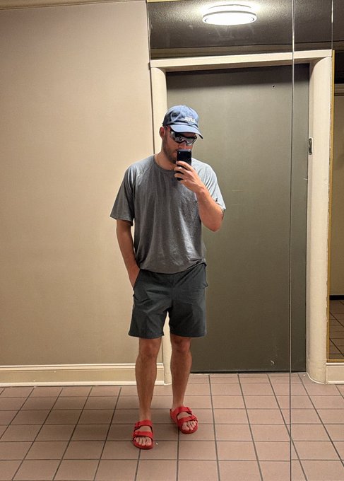 A mirror selfie of a man wearing a blue cap, glasses, a gray t-shirt, gray shorts, and red sandals, standing in what appears to be a public restroom.