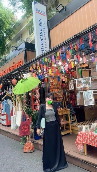 A woman wearing a green face mask and a black outfit stands in front of 'Chokchai 4Handcraft' shop, with a banner for 'BATHHOA SEN SEN University' visible above and colorful lanterns hanging around.