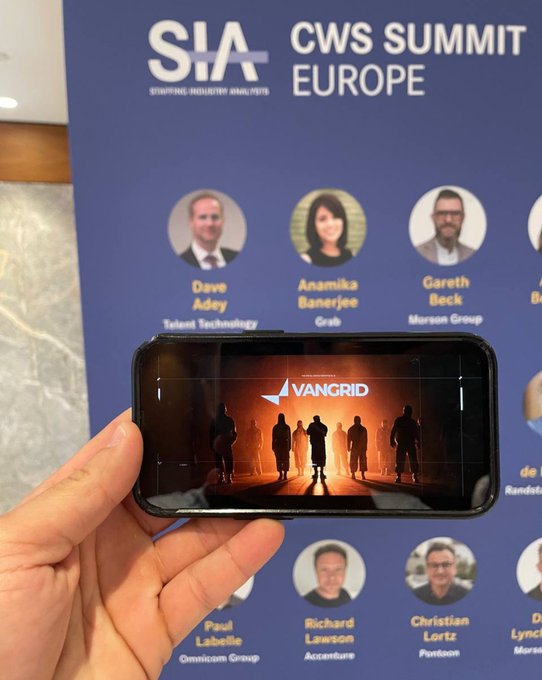 A hand holds a smartphone displaying a VanGrid logo over silhouettes, in front of a poster for the 'CWS SUMMIT EUROPE' featuring speakers Dave Adey, Anamika Banerjee, Gareth Beck, Paul Labelle, Richard Lawson, and Christian Lortz.