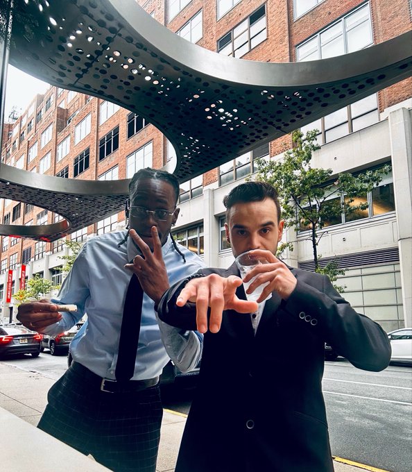 Two young men posing playfully on a city street, one wearing glasses and a white shirt, the other in a dark suit holding a cup, photographed from a low angle beneath a curved architectural structure.