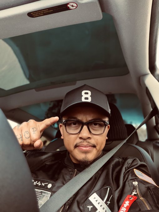 A man wearing a black cap with the number "8" and a leather jacket sits in a car, flashing a "HOPE" hand gesture tattoo toward the camera. His jacket features American flag patches and text reading "TAKEO" and "W4X." The casual, confident pose matches the request for a Congress-style pose.