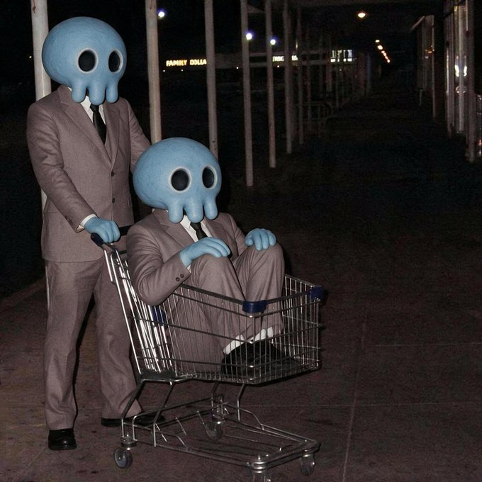 Two figures wearing grey suits with blue octopus-like masks, one pushing the other who is seated inside a metal shopping cart on a dark street at night.