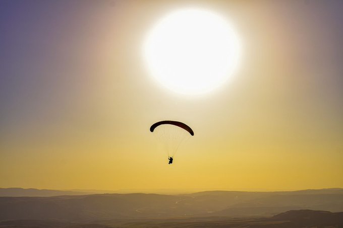 A scenic photograph of a paraglider silhouetted against a bright, oversized sun in a golden-hued sky over distant mountains.