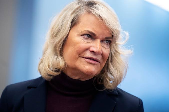 Portrait of a woman with shoulder-length blonde hair wearing a dark turtleneck and blazer against a blue background.