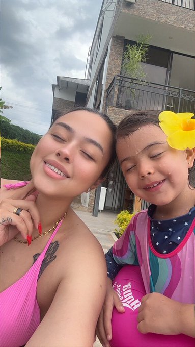 A selfie of a smiling woman in a pink top and a young girl wearing a colorful swimsuit, both outdoors in front of a residential building.
