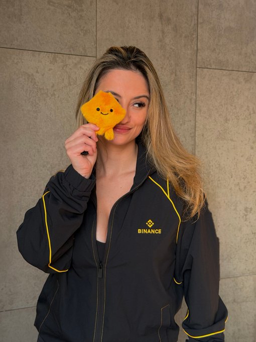 A blonde woman wearing a black Binance branded jacket holds a yellow star-shaped plush toy over one eye.