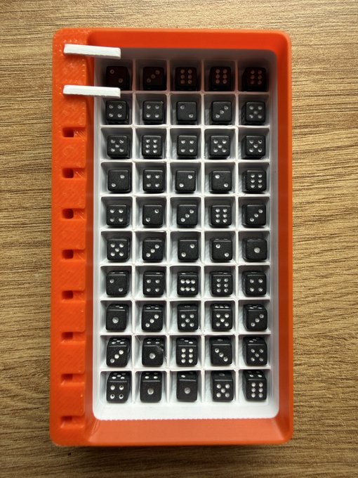 A 3D-printed orange and white tray containing numerous black dice with white dots arranged in a grid pattern on a wooden surface.