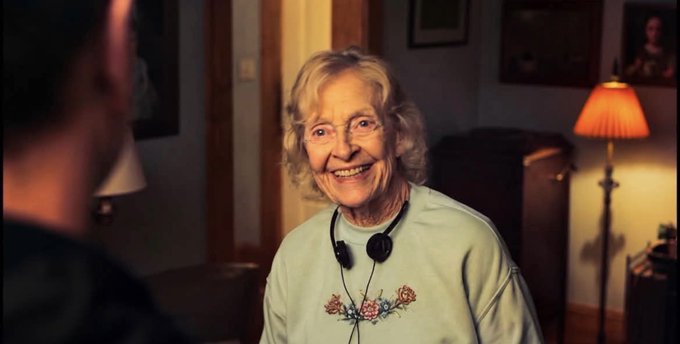 An elderly woman with short blonde hair smiles warmly while wearing headphones around her neck, appearing to be in conversation with someone across from her.